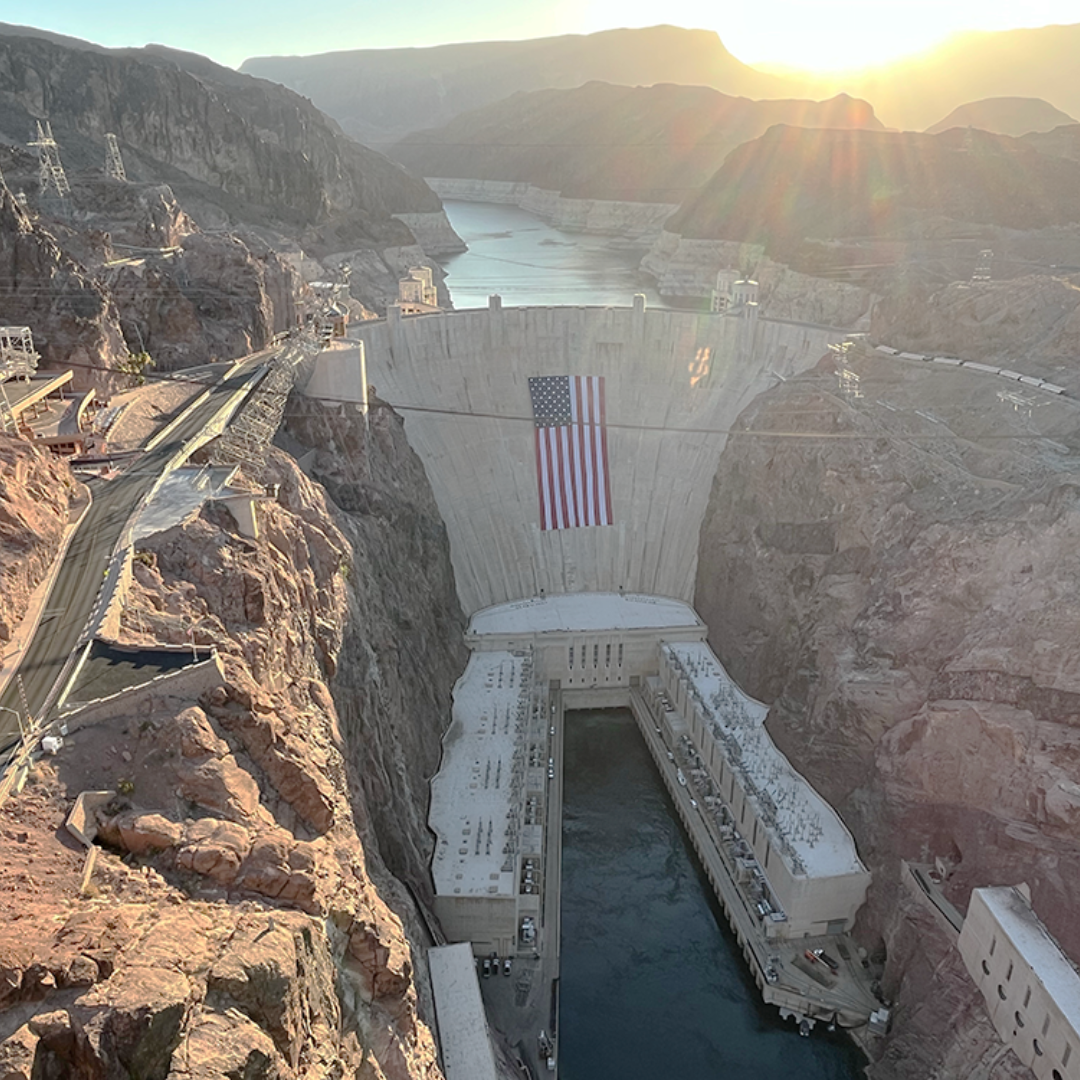 Hoover Dam at golden hour, the American flag draped down its concrete face, transmission towers on the canyon rim.